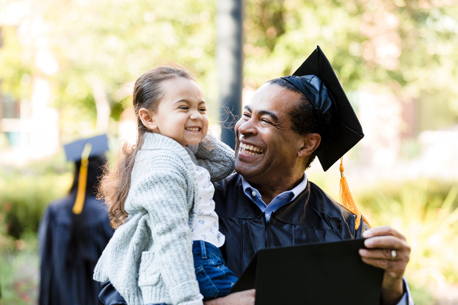 A graduate in cap and gown laughing with his young daughter